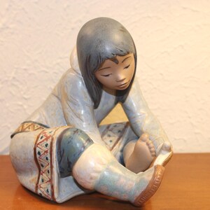 May include: A porcelain figurine of a young girl wearing a blue and white patterned dress, sitting with her legs crossed and looking down.