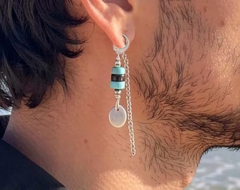 May include: A silver hoop earring featuring a dangling chain, a small round silver charm, and a cylindrical bead with horizontal stripes in black and turquoise blue. The earring is secured through the earlobe with a polished silver hoop component.