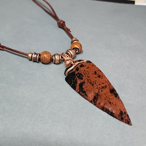 May include: A necklace featuring a brown and black speckled arrowhead pendant wrapped in copper wire. The pendant hangs from a brown leather cord accented with metallic and speckled brown beads, secured by a sliding knot mechanism for adjustment.