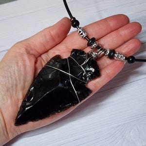 May include: A black obsidian arrowhead pendant necklace. The arrowhead is wrapped in silver wire and hangs from a black cord necklace. The necklace features decorative silver beads and black beads.