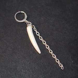 May include: A silver-toned hoop earring featuring a dangling white, curved, horn-shaped pendant and a separate silver-toned chain link strand attached to the same hoop, creating a layered, asymmetrical design with metallic and smooth white elements.