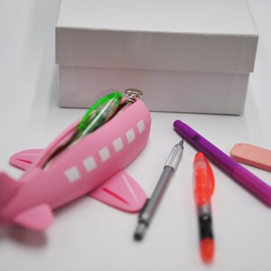 May include: Pink airplane-shaped pencil case with a zipper, filled with colorful pens and pencils. A white box is in the background.