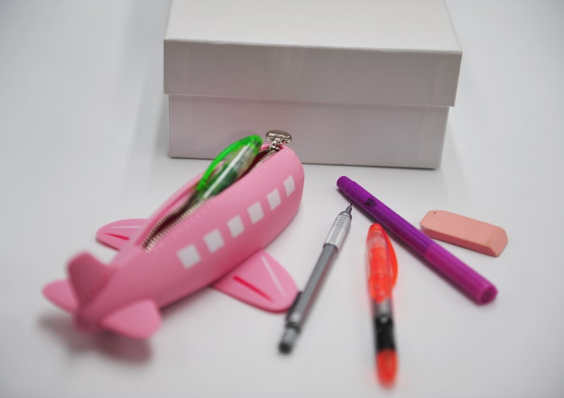May include: Pink airplane-shaped pencil case with a zipper, filled with colorful pens and pencils. A white box is in the background.