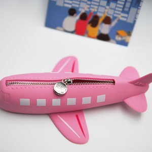 May include: A pink airplane-shaped pencil case with a zipper closure and a silver zipper pull. The case has white rectangular windows and a small silver tag with the word "FLY" on it.