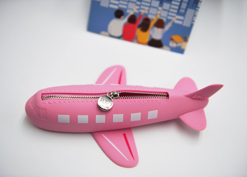 May include: A pink airplane-shaped pencil case with a zipper closure and a silver zipper pull. The case has white rectangular windows and a small silver tag with the word "FLY" on it.