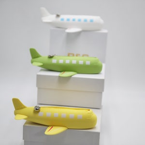 May include: Three airplane-shaped zippered pouches, one white, one green, and one yellow, stacked on top of each other on white boxes.