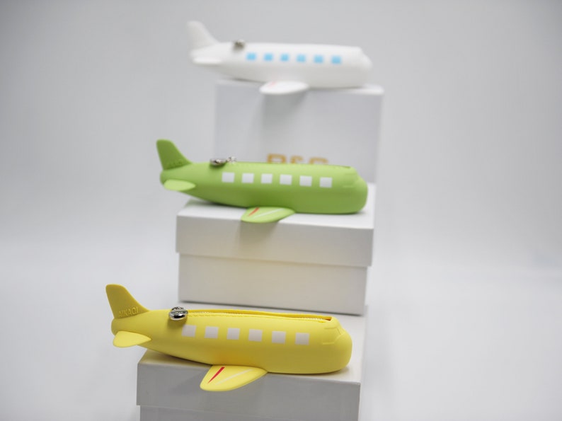May include: Three airplane-shaped zippered pouches, one white, one green, and one yellow, stacked on top of each other on white boxes.