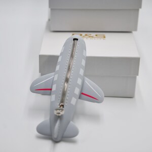 May include: A gray airplane-shaped zippered pouch with a white zipper and red accents. The pouch is sitting on a white surface.