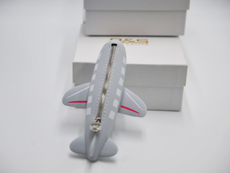 May include: A gray airplane-shaped zippered pouch with a white zipper and red accents. The pouch is sitting on a white surface.