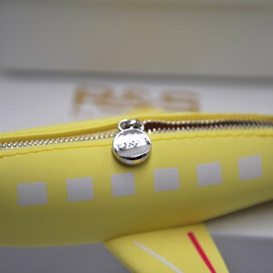 May include: Yellow airplane-shaped zippered pouch with a silver zipper pull. The pouch has white squares on the side and a red stripe on the bottom.