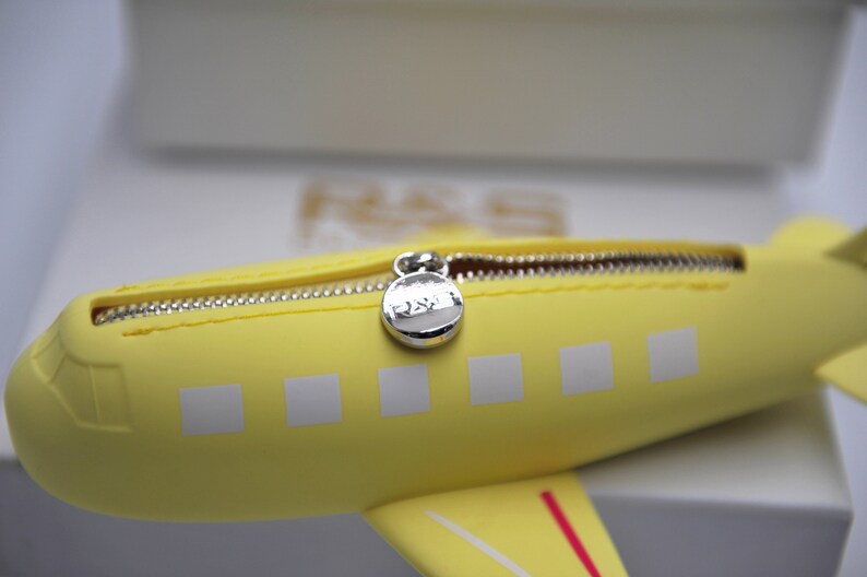 May include: Yellow airplane-shaped zippered pouch with a silver zipper pull. The pouch has white squares on the side and a red stripe on the bottom.
