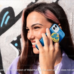 May include: A woman with pink highlights in her hair is talking on a blue smartphone. The phone has a yellow and blue pop socket with a bird design and the word "Moscow" written on it.