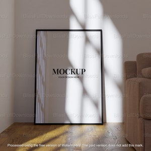 May include: A black framed mockup poster with the text "MOCKUP YOUR DESIGN HERE" on a white background. The poster is leaning against a white wall with a brown couch in the background. Sunlight is streaming through a window, casting shadows on the wall and floor.