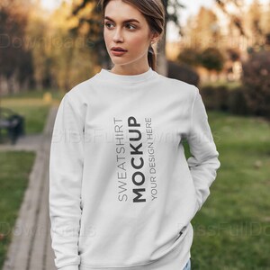 May include: A white sweatshirt mockup with the text "SWEATSHIRT MOCKUP YOUR DESIGN HERE" in black. The sweatshirt is worn by a person in blue jeans, standing outside on a path. The background is blurred with trees and foliage.