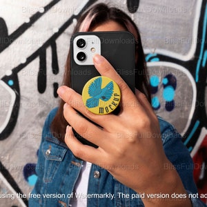 May include: A person is holding a black smartphone with a yellow and blue phone grip. The phone grip has a blue bird design and the words "MOCKUP" in white letters. The person is standing in front of a graffiti wall.