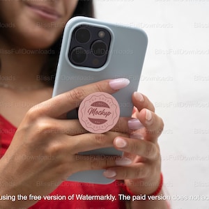 May include: A person is holding a light blue smartphone with a pink pop socket attached to the back. The pop socket has a white circle with the text "POP SOCKET MockUp POP SOCKET" in brown lettering.