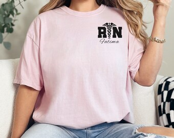 May include: A pink t-shirt with a black graphic of a medical caduceus and the letters "RN" with the name "Fatima" underneath.