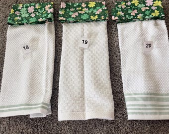 May include: Three white kitchen towels with green and floral patterned tops. Each towel has a different texture and a number on a small white tag. The tops feature green fabric with pink, yellow, and green shamrocks.