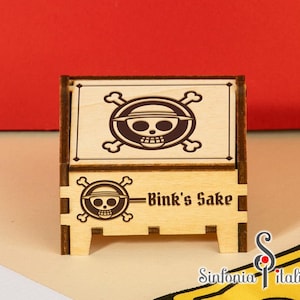 May include: A wooden box with a pirate-themed design. The box features a skull and crossbones logo with a straw hat. The text "Bink's Sake" is displayed on the front. The box is made of light-colored wood.