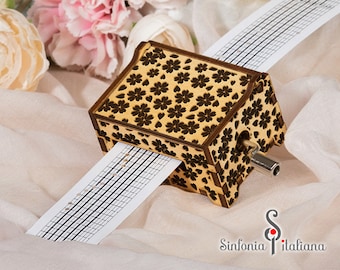 May include: A wooden music box with a floral design and a paper music sheet. The music box has a crank on the side to wind it up.