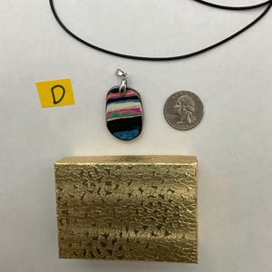 May include: A colorful, abstract pendant with a silver bail hangs on a black cord. The pendant features horizontal stripes of pink, blue, black, and white. A gold textured box and a quarter are also in the image.