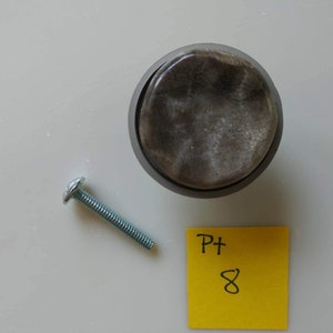 May include: A round, gray, marble-like cabinet knob with a silver screw. The knob is on a white surface with a yellow sticky note that says "Pt 8".