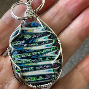 May include: Oval pendant with a colorful, swirled design in shades of black, blue, green, and white, encased in a silver wire frame. The pendant has a unique, abstract pattern and is suspended from a silver wire bail.
