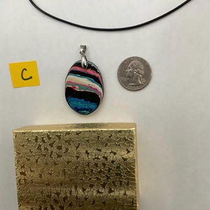 May include: Oval pendant necklace with a colorful, layered design in shades of black, blue, pink, and white. The pendant is suspended from a black cord. A gold textured gift box and a quarter are also in the image.