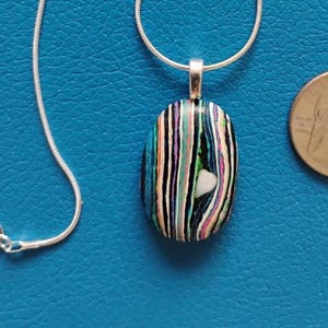May include: A silver necklace with a pendant featuring a colorful, striped design. The pendant is an oval shape with vertical stripes of blue, black, white, pink, and green. A U.S. quarter is next to the pendant for size comparison.