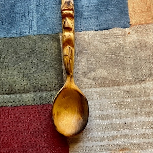 May include: A wooden spoon with a carved face on the handle. The spoon is brown and has a natural finish. It is lying on a multi-colored fabric background.