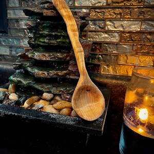 Può includere: Un cucchiaio di legno con una finitura in legno naturale. Il cucchiaio è appoggiato su una fontana di roccia con piccoli sassi e una candela accesa sullo sfondo.