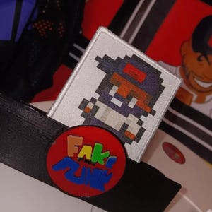 May include: A white embroidered patch with a pixelated image of a person wearing a black hat and white shirt. The patch is attached to a black plastic object with a red, yellow, blue, and green logo that says "FAKE FUNK".