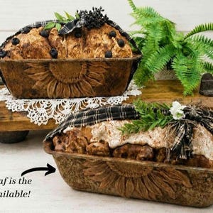 May include: Two rectangular metal loaf pans with embossed sunflower designs on the sides. The front pan contains a baked loaf topped with white frosting and a black plaid ribbon, with text stating: This loaf is the one available!