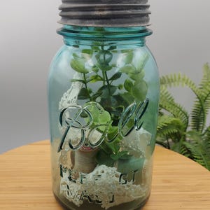 May include: A blue Ball mason jar with a dark metal lid, filled with greenery, lace, and spools of thread. The jar has the Ball logo embossed on the front. The jar is sitting on a wooden surface.