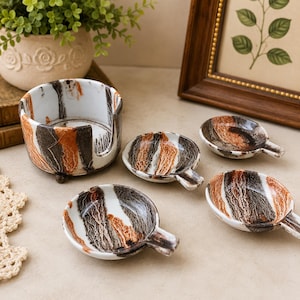 May include: A ceramic set featuring a cylindrical holder with a side cutout and four small, round, handled dishes. Each piece displays a marbled pattern of white, dark brown, and burnt orange, with a glossy finish and a distinct textured appearance.
