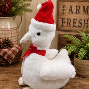 May include: A plush white goose figurine featuring a soft, fuzzy texture, wearing a bright red Santa hat with a white pom-pom and a matching red velvet ribbon tied around its neck. The goose has a white beak and feet with a subtle grey snowflake pattern.