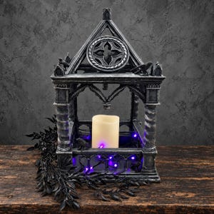 May include: A gray, weathered-stone-look lantern featuring a peaked roof with a circular quatrefoil window, four spiraled columns, and two small raven figures on the roof. Inside, a cream-colored pillar candle sits amidst purple string lights.