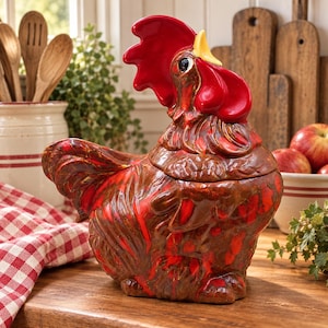 May include: A ceramic rooster-shaped cookie jar featuring a glossy finish with deep brown and vibrant red color variations. The rooster has a prominent red comb, a yellow beak, and a textured plumage design covering the entire body.