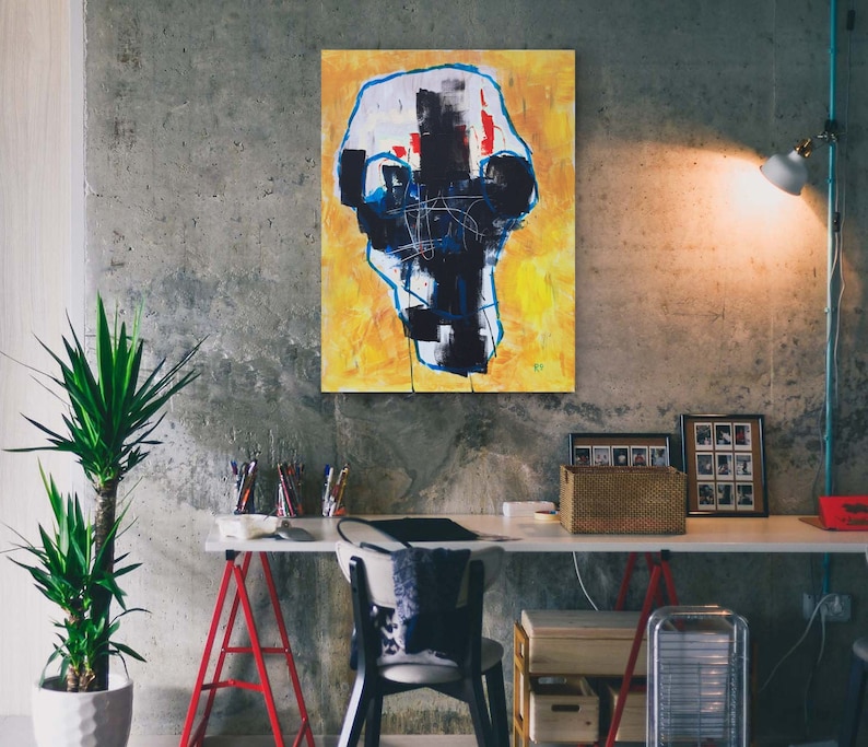 May include: Abstract art piece featuring a skull-like shape with black, white, red, and blue brushstrokes against a yellow background. The artwork is displayed on a concrete wall in a room with a desk and a plant.