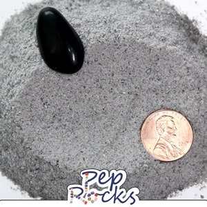 May include: A close-up of a small pile of gray sand with a black stone and a US penny on top. The sand is fine-grained and has a slightly sparkly appearance.