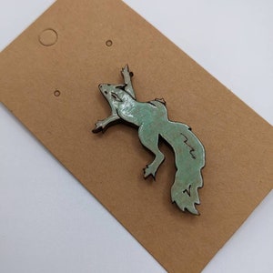 Puede incluir: Broche en forma de ardilla de color turquesa con acabado envejecido. El broche es de metal y tiene un diseño detallado. Se muestra en una etiqueta de cartón marrón con dos pequeños agujeros.
