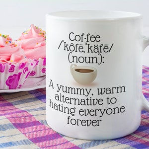 May include: White coffee mug with the text "Coffee /k&ocirc;fē, k&auml;fē/ (noun): A yummy, warm alternative to hating everyone forever." Pink frosted cupcakes with sprinkles sit on a plate next to the mug.