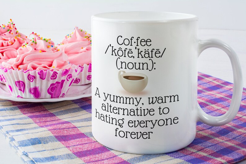 May include: White coffee mug with the text "Coffee /k&ocirc;fē, k&auml;fē/ (noun): A yummy, warm alternative to hating everyone forever." Pink frosted cupcakes with sprinkles sit on a plate next to the mug.
