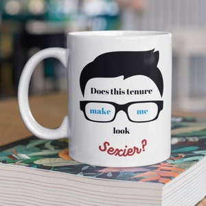 May include: White ceramic mug with a black silhouette of a person's hair and glasses. The text on the mug reads "Does this tenure make me look Sexier?" in black, blue, and red lettering. The mug has a white handle.