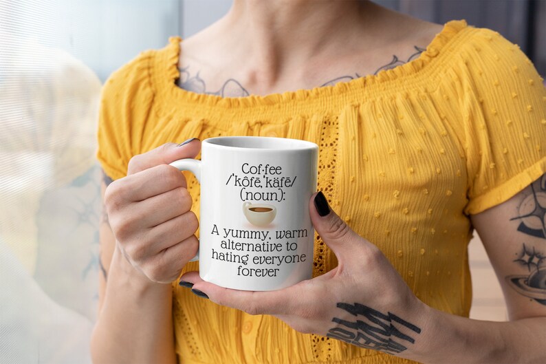 May include: A white ceramic coffee mug with the text "Coffee /'kōfē, kāfē/ (noun): A yummy, warm alternative to hating everyone forever." The mug has a handle and a small coffee cup graphic.