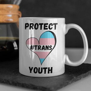 May include: White ceramic mug with the words "PROTECT YOUTH" in black, framing a heart-shaped design. The heart features the transgender pride flag colors and the hashtag "#TRANS". The mug has a curved handle.