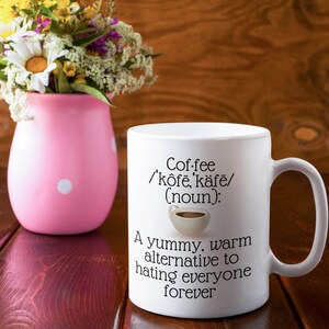 May include: White coffee mug with the definition of coffee printed on it, including a small coffee cup graphic. The text reads "A yummy, warm alternative to hating everyone forever." A pink vase with white polka dots and a bouquet of flowers is next to the mug.