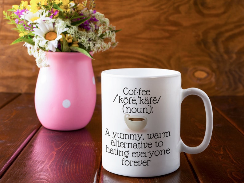 May include: White coffee mug with the definition of coffee printed on it, including a small coffee cup graphic. The text reads "A yummy, warm alternative to hating everyone forever." A pink vase with white polka dots and a bouquet of flowers is next to the mug.