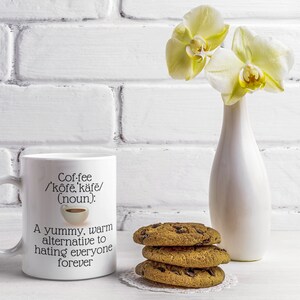 May include: A white coffee mug with the text "Coffee /'k&ocirc;fe, k&auml;fe/ (noun): A yummy, warm alternative to hating everyone forever." Next to the mug are three chocolate chip cookies and a white vase with yellow orchids.