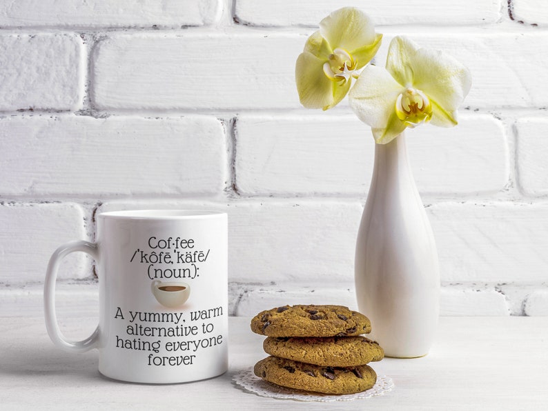 May include: A white coffee mug with the text "Coffee /'k&ocirc;fe, k&auml;fe/ (noun): A yummy, warm alternative to hating everyone forever." Next to the mug are three chocolate chip cookies and a white vase with yellow orchids.